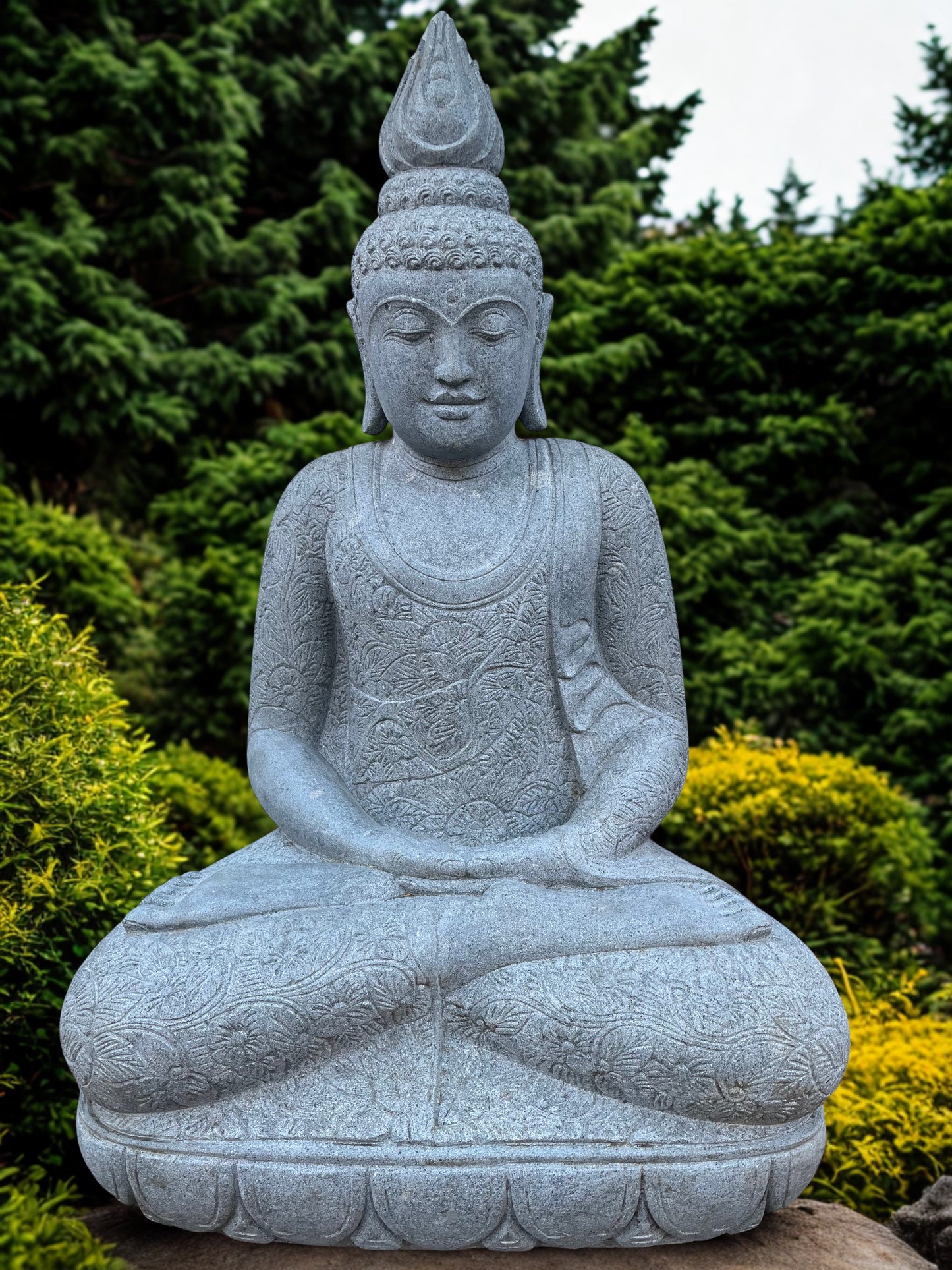 Stone statue of a seated Buddha in a garden setting with greenery.