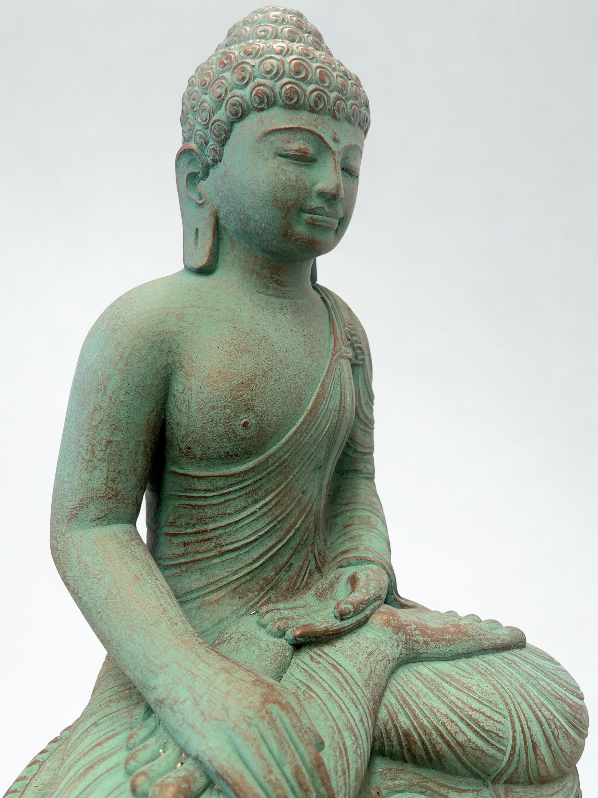 A seated Buddha statue with a weathered green patina and reddish-brown undertones, shown against a plain light gray background.