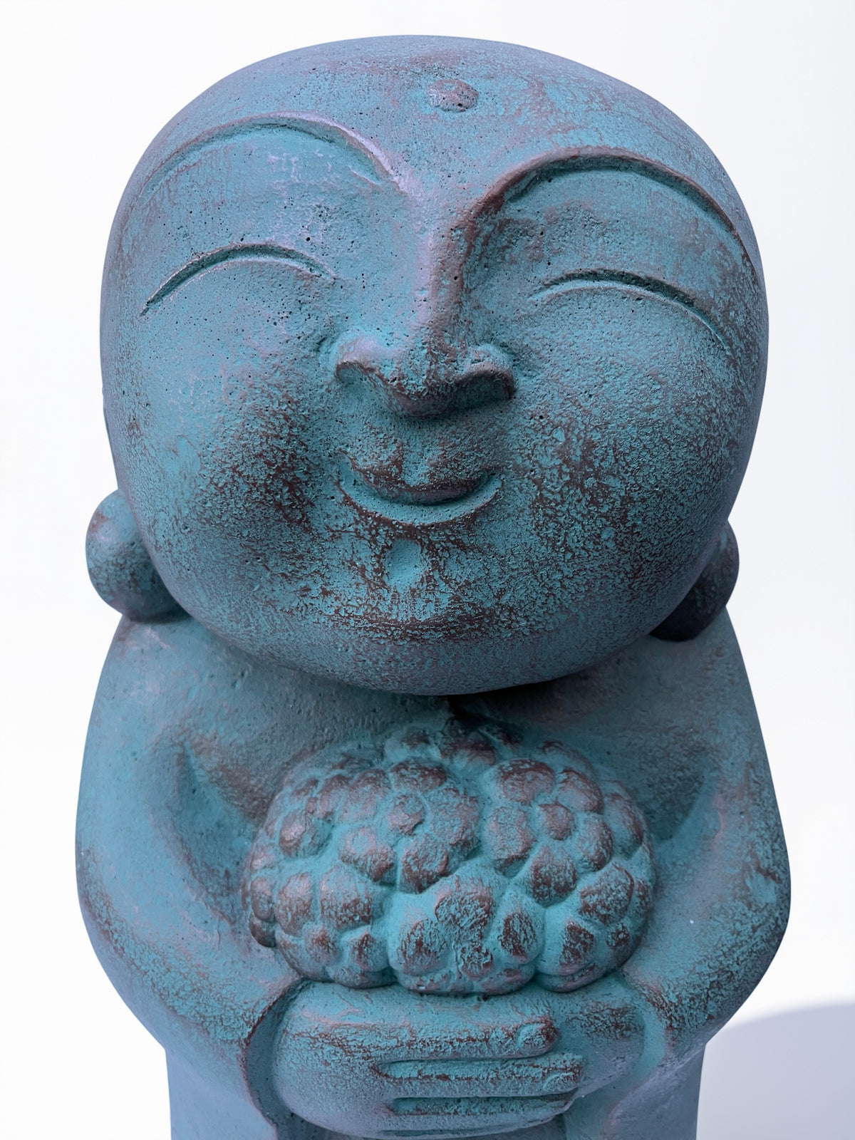 A close-up of a teal-blue stone concrete Jizo figurine holding a cluster of flowers, set against a plain white background.