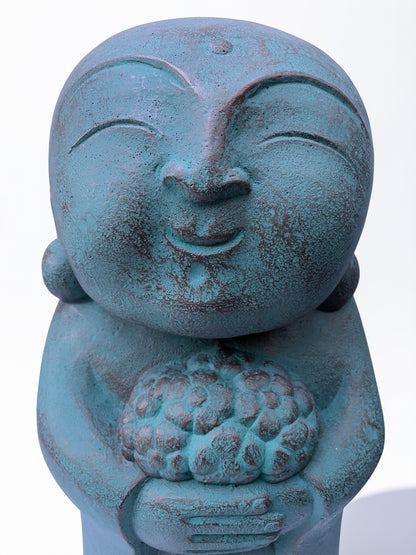 A close-up of a teal-blue stone concrete Jizo figurine holding a cluster of flowers, set against a plain white background.