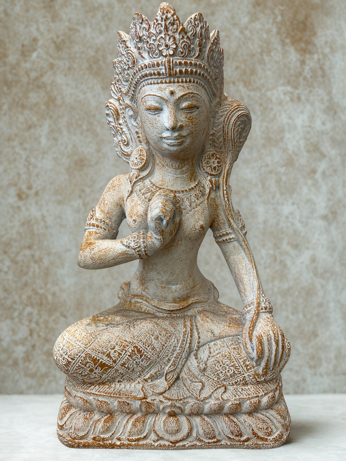 An intricately adorned beige stone statue of a seated female deity with ornate jewelry and a detailed crown, set against a textured neutral background.