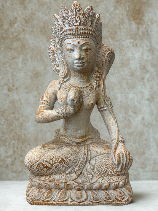 An intricately adorned beige stone statue of a seated female deity with ornate jewelry and a detailed crown, set against a textured neutral background.