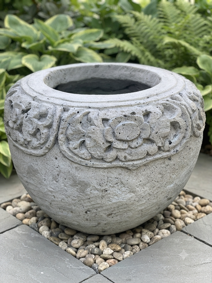 A round concrete garden planter with carved floral patterns around the upper section, placed outdoors on small pebbles with green foliage in the background.