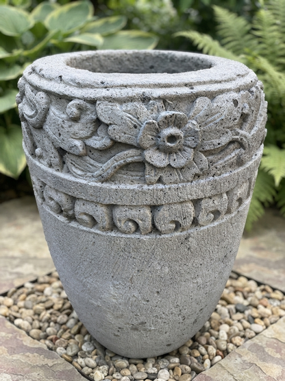 A gray stone garden planter with carved floral and scroll patterns, displayed outdoors on a bed of small pebbles with green foliage in the background.