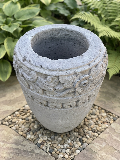 A gray concrete planter pot with carved floral and scroll patterns, placed on a bed of small pebbles with green garden foliage in the background.