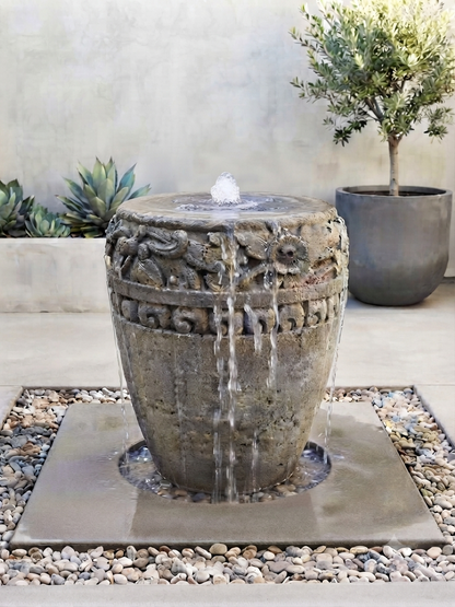 A round stone fountain with carved floral details and a small pool of water at the top, set outdoors on a square stone base surrounded by small pebbles. A potted tree and succulents are visible in the background against a light stucco wall.