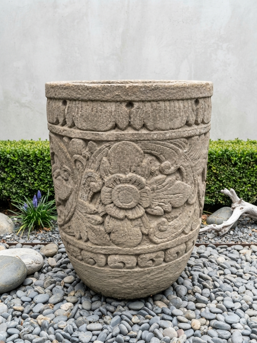 A large round stone planter pot with carved floral and scroll patterns, placed on gray pebbles with green hedges and a light gray wall in the background.