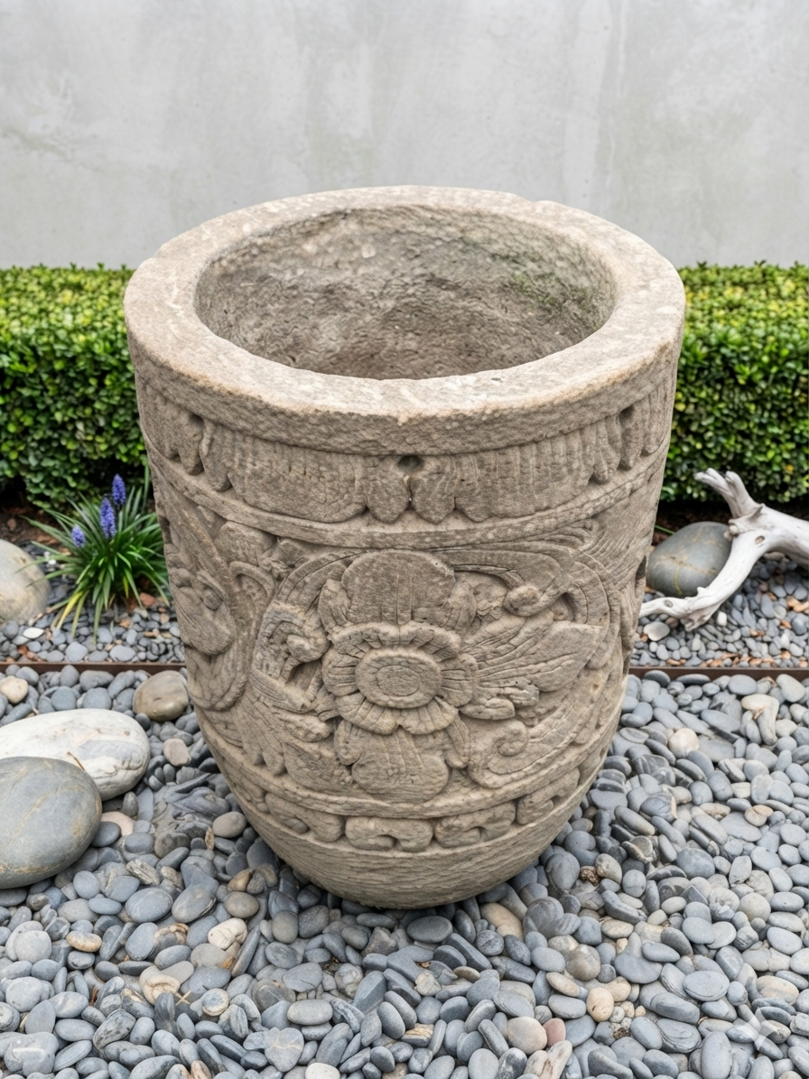 A large round stone planter pot with carved floral patterns, placed on a bed of gray pebbles outdoors with green hedges and a concrete wall in the background.