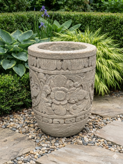 A large round stone planter with carved floral and scroll patterns, placed outdoors on a stone path surrounded by gravel, green shrubs, and plants.