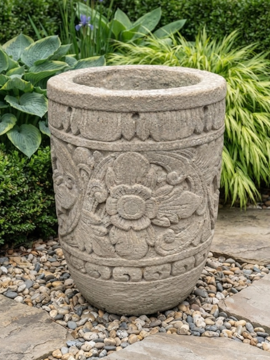 A cylindrical stone planter with carved floral and scroll patterns, placed outdoors on a bed of small gray and beige pebbles with green garden foliage in the background.