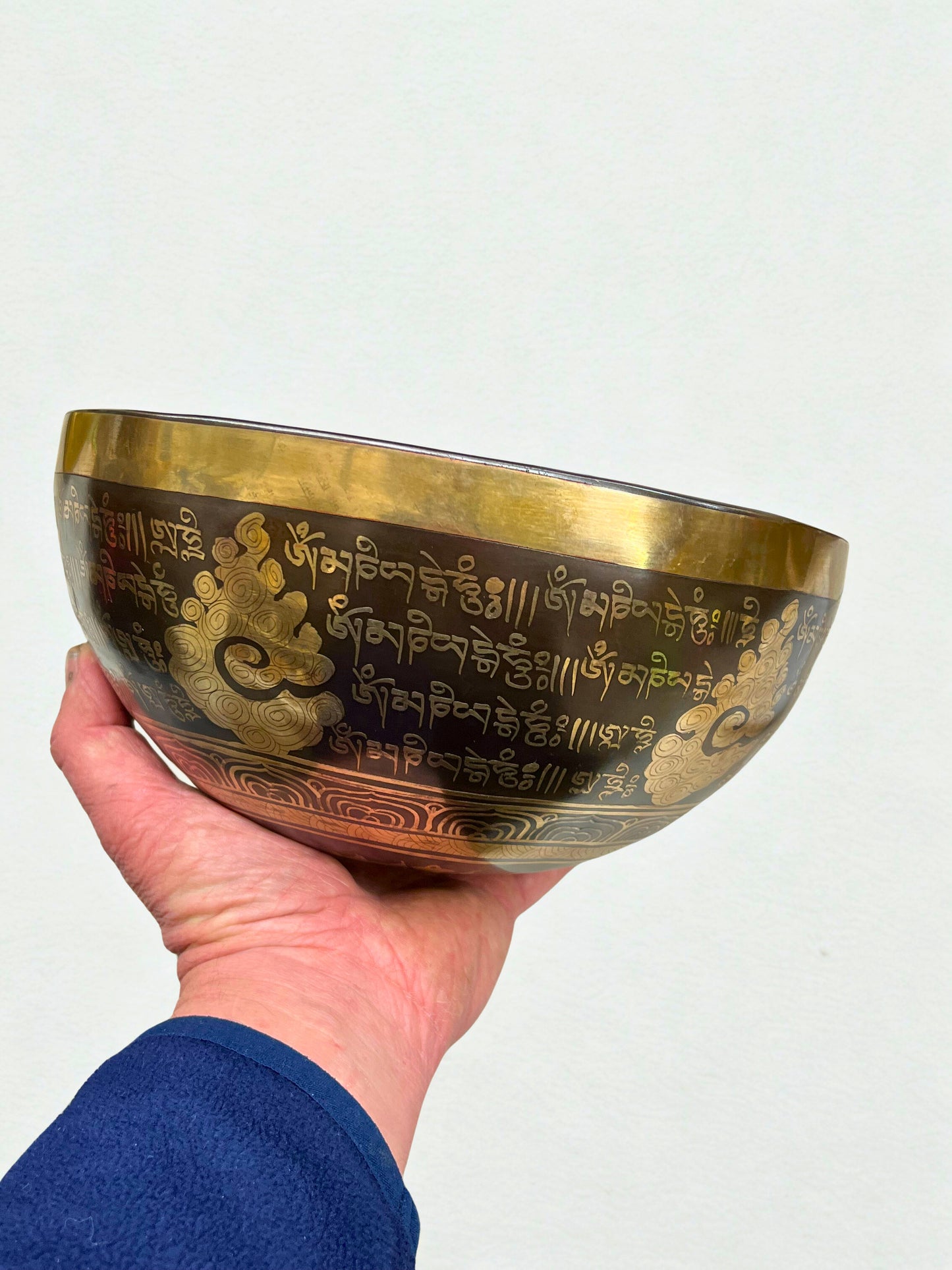 Hand holding a brass singing bowl with engraved text and patterns on a light gray background